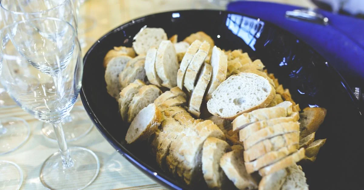 Baguette tranchée servie dans un bol noir avec des verres à vin sur une table en bois.