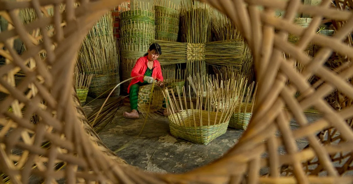Une femme tisse habilement des paniers en bambou à l'intérieur d'un atelier, un aperçu à travers un grand cadre de panier.