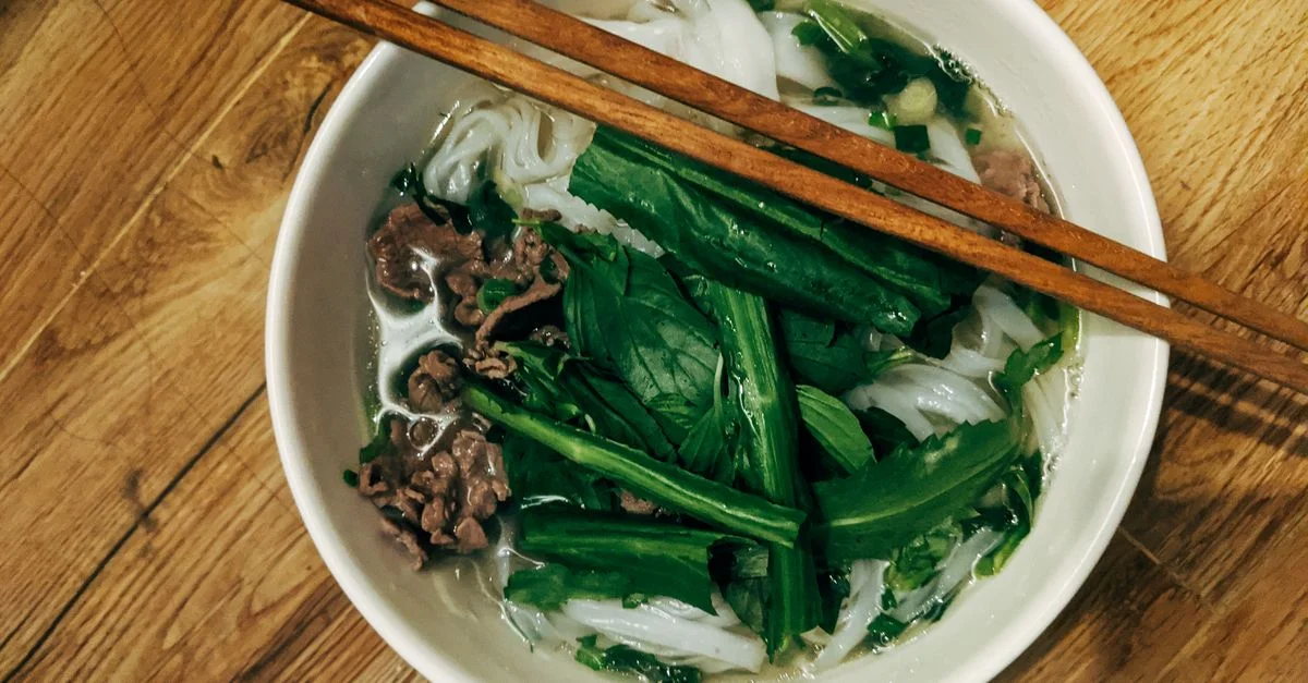 Vue de dessus d'un bol de pho vietnamien avec des herbes fraîches et des baguettes sur une table en bois.
