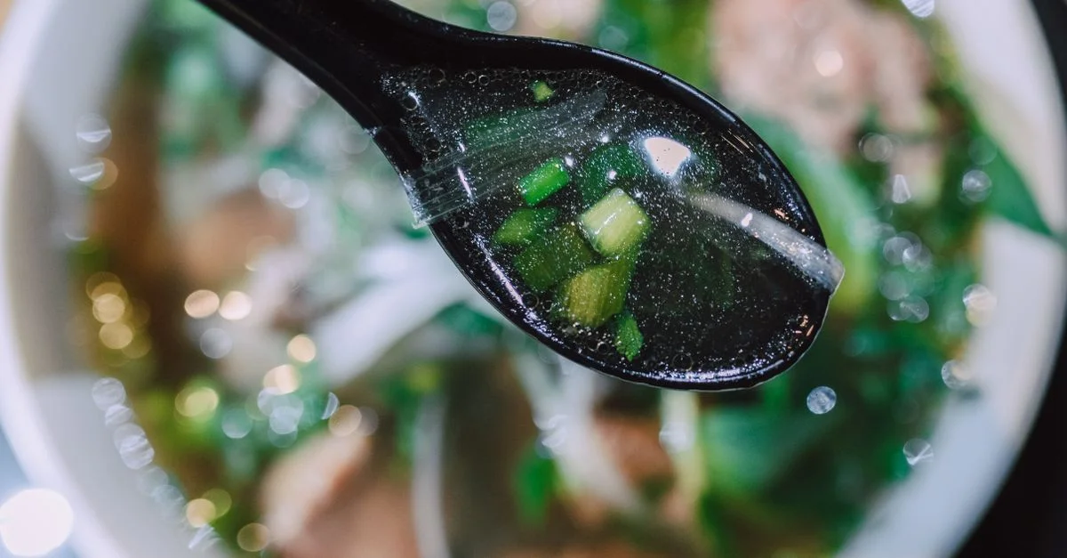 Gros plan appétissant de soupe pho vietnamienne avec des oignons verts sur une cuillère noire, mettant en valeur des saveurs riches.