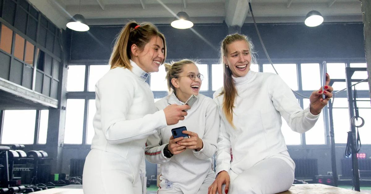 Trois femmes escrimeuses en uniformes blancs riant et prenant un selfie à l'intérieur.