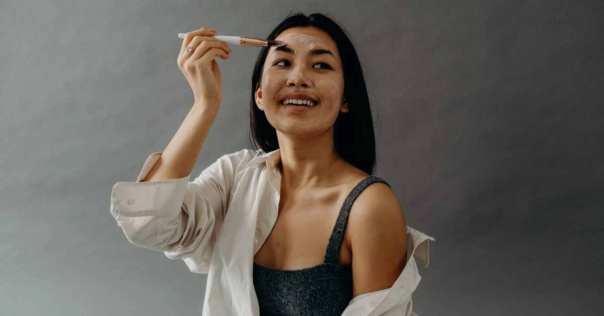 Femme asiatique souriante appliquant une crème faciale avec un pinceau, promouvant la beauté et les soins de la peau dans un cadre de studio.