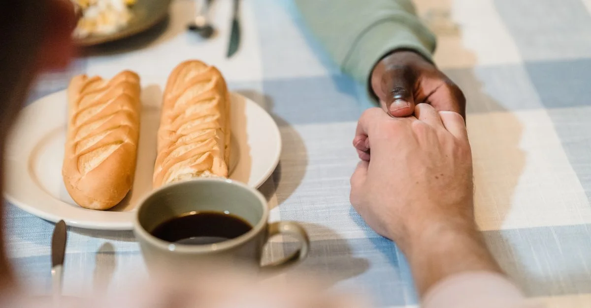 Un couple se tient par la main au-dessus d'une table de petit-déjeuner avec du pain et du café, symbolisant la connexion et la convivialité.